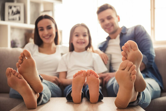 Charmantes kleines Mädchen und ihre schönen jungen Eltern schauen in die Kamera und lächeln, während sie auf dem Sofa zu Hause sitzen. Nackte Füße im Fokus.